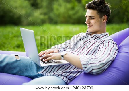 Handsome Young Man Lying On Inflatable Sofa Lamzac Play Games On Laptop While Resting On Grass In Pa