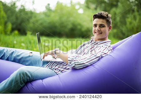 Handsome Young Smile Man Lying On Inflatable Sofa Working On Laptop While Resting On Grass In Park