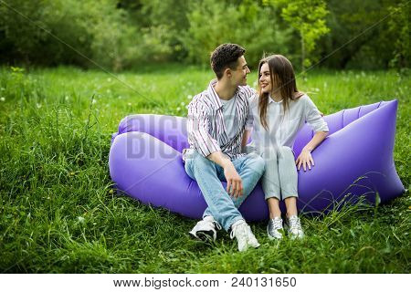 Love Couple Lying On Inflatable Sofa Lamzac On Laptop While Resting On Grass In Park Looking Each Ot