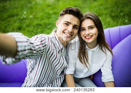 Young Couple Sitting On Inflatable Sofa Lamzac And Take Selfie On Phone While Resting On Grass In Pa