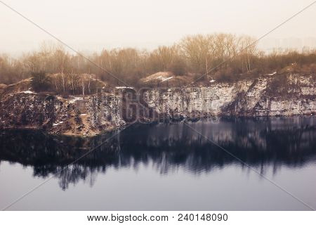 Fantastic Autumn Landscape With Dark Blue Lake In The Quarry And Orange Sky And Trees