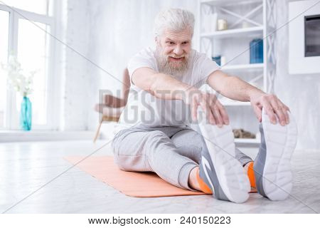 Supple Muscles. Cheerful Elderly Man Sitting On The Yoga Mat And Touching His Toes While Stretching 