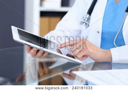 Woman Doctor Using Tablet Computer While Sitting At The Glass Table In Hospital Office, Closeup. Hea