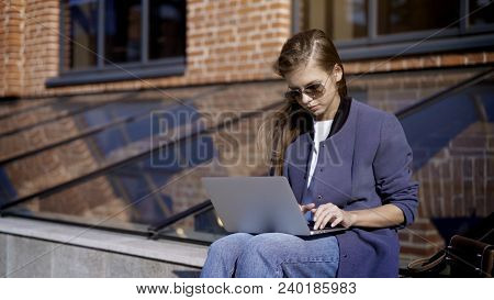 A Young Pretty Concentrated Girl In Jeans In Sunglasses Is Working With A Laptop Outisde Sitting On 