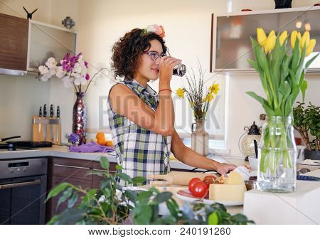 Brunette Female With Curly Hair Drinks Water In A Kitchen.