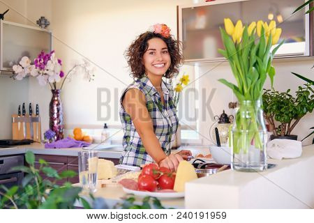 Positive Brunette Female With Curly Hair Makes Salad With Tomatoes And Potato In A Home Kitchen.
