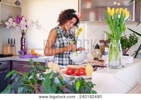 Positive Brunette Female Makes Dough In A Kitchen With Yellow Tulips On Background.