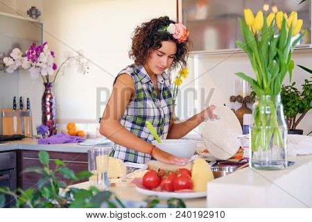 Positive Brunette Female Makes Dough In A Kitchen With Yellow Tulips On Background.