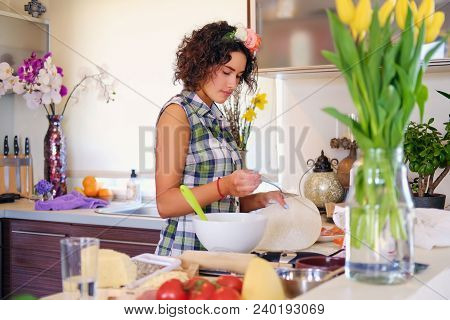 Positive Brunette Female Makes Dough In A Kitchen With Yellow Tulips On Background.