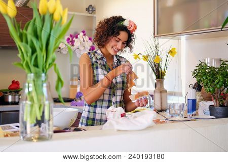 Positive Brunette Female Makes Dough In A Kitchen With Yellow Tulips On Background.