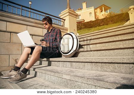 The Guy Is Sitting On The Street On The Stairs With A Laptop And A Mono Wheel