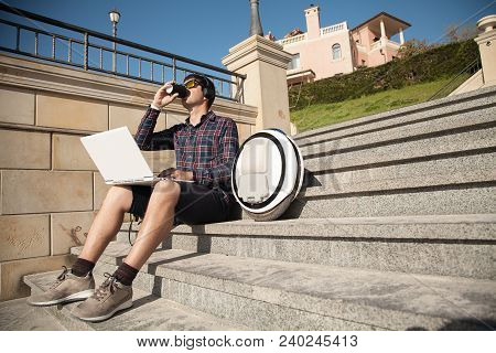 The Guy Is Sitting On The Street On The Stairs With A Laptop And A Mono Wheel