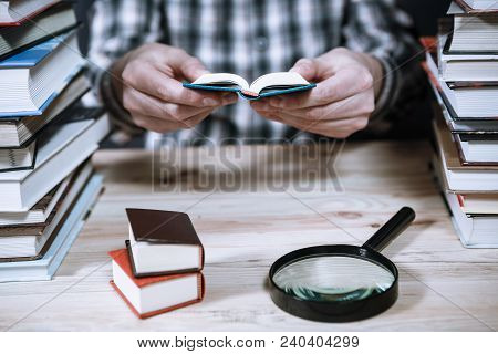 A Man Reading A Miniature Book Among The Big Books In The Library At The Wooden Table.