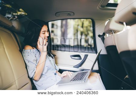 Attractive Young Business Woman Working On Her Laptop At The Back Sit Of A Car And Talking On The Ph