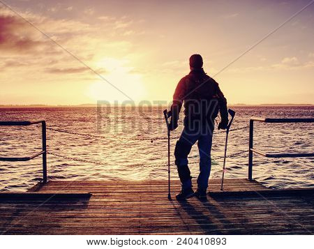 Hurt Man With Hooded Jacket And Forearm Crutches  Looking Sadly Into Sea Water. Traveler  Stand On S