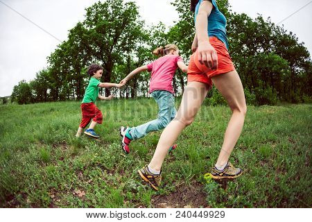 A Woman With Children Runs Along The Green Grass. Mom Holds Her Sons By The Hands And Laughs. A Boy 