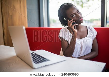 Portrait Of African American Girl Sitting At Cafe And Working On Her Laptop Talking On Her Cellphone