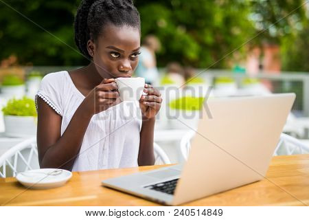Portrait Of African American Girl In Glasses Sitting At Cafe With Laptop And Holding Cup Of Coffee I
