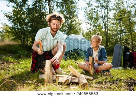 Happy little boyscout looking at his father preparing firewood for campfire on sunny day