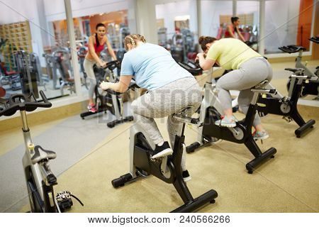 Two plus-sized girls sitting on bicycles and training hard while trainer instructing them in gym