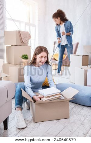 Fruitful Cooperation. Charming Female Student Unpacking A Pile Of Books While Her Roommate Cleaning 
