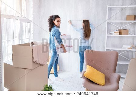 Helping Her Roommate. Pretty Curly-haired Girl Posing With A Picture In Her Hands While Waiting For 