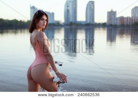 Young Woman In A Swimsuit Stands On The River Bank With A Bicycle.