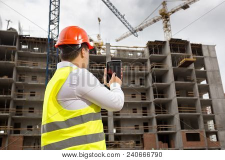 Rera View Image Of Businessman In Safety Vest And Hardhat Standing On Building Site And Using Digita