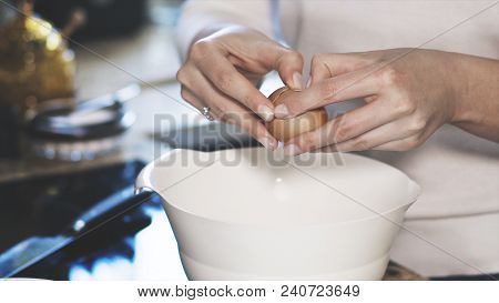A Young Woman Dressed In A White Shirt Is Breaking Up A Brown Egg Into A White Bowl. Closeup Of Hand