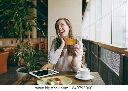 Woman Sitting Near Big Window In Coffee Shop At Table With Credit Card, Cup Of Coffee, Cake, Relaxin