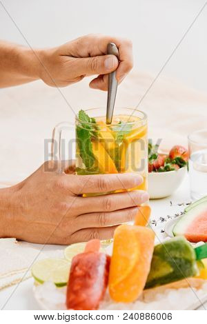 young man stirring a jar with a refreshing drink, with orange slices and mint, placed on a table next to a bowl with strawberries, a plate with watermelon and a plate with different homemade ice pops