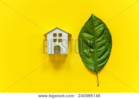 Miniature Toy Model House With Green Leaves On Yellow Colourful Trendy Backgdrop. Eco Village, Abstr