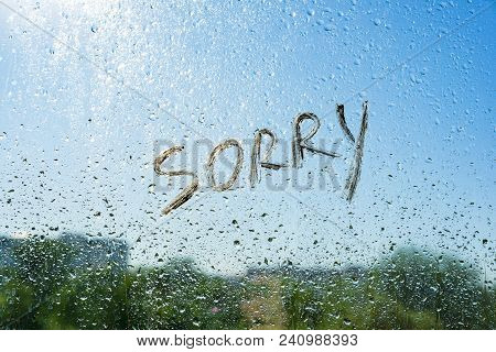 Word Sorry On The Window With Raindrops. Background Drop Of Water On The Glass, Blue Sky, Sun, City