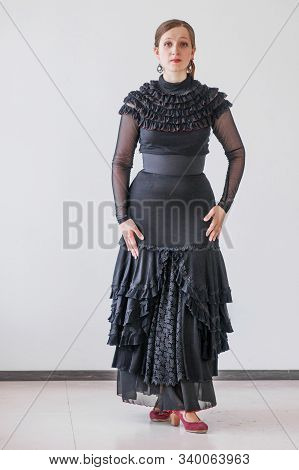 Beautiful Young Woman In A Black Elegant Dress And Dancing Flamenco On A Background Of A Gray Wall. 