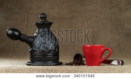 A Steel Measuring Spoon With Roasted Coffee Beans And A Made Of Black Clay сoffeepot With Red Cup On