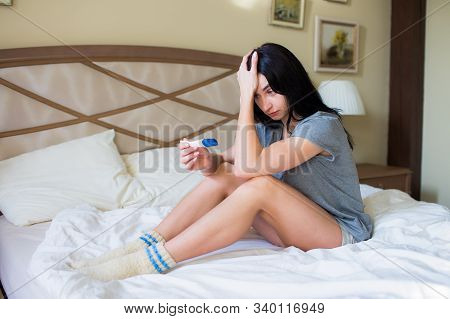 Frustrated Young Brunette Woman With Pregnancy Test Sitting On A Bed. Negative Pregnancy Test.