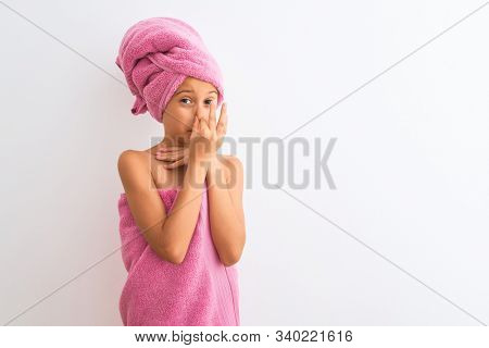 Beautiful child girl wearing shower towel after bath standing over isolated white background smelling something stinky and disgusting, intolerable smell, holding breath with fingers on nose. Bad smell