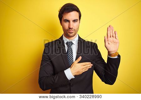 Young handsome businessman wearing suit and tie standing over isolated yellow background Swearing with hand on chest and open palm, making a loyalty promise oath