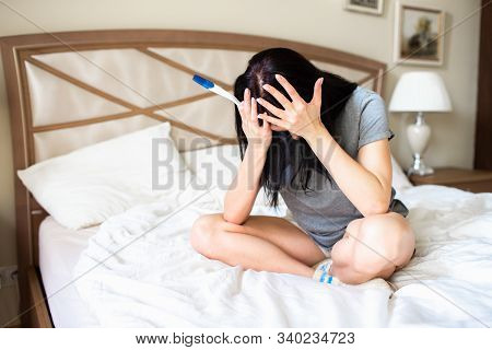 Frustrated Young Brunette Woman With Pregnancy Test Sitting On A Bed. Negative Pregnancy Test.