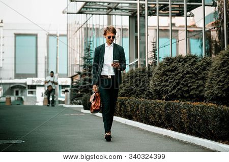 The Well-dressed Businessmen Is Walking Down The Green City Street And Reading Something In His Mobi