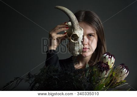 Portrait Of A Young Dark-haired Girl Holding Half A Goat Skull In Front Of Her Face And A Bouquet Of
