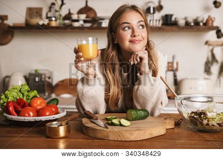 Image of pleased cute woman wearing casual clothes drinking juice while making lunch in cozy kitchen