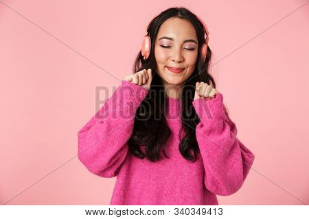 Image of optimistic beautiful asian girl with long dark hair smiling and listening to music with headphones isolated over pink background