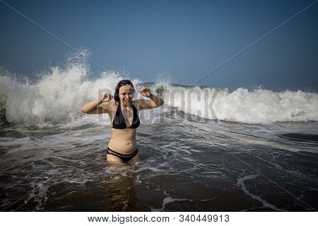 A Young Woman In A Black Swimsuit Spends Time In The Sea Or Ocean. The Girl Takes Refuge From The Wa