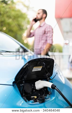 Handsome Young Guy With A Beard Talking On The Phone Near His Electric Car.