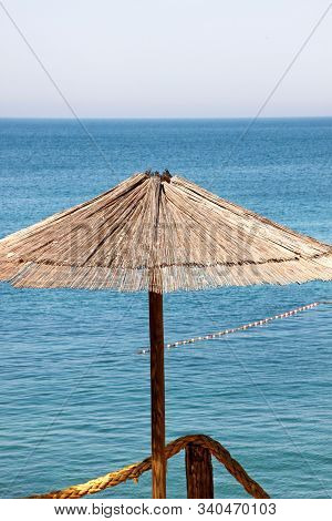 Lonely Wicker Sun Umbrella At Mediterranean Beach By Sea. Natural Bamboo Sunshades And Summer Umbrel