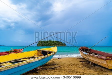 Colorful old wooden fishing boats docked by water on a beautiful beach coast. White sand sea shore landscape on tropical Caribbean island. Holiday weekend/ summer vacation setting at Pagee / Pajee Beach in Port Maria, Jamaica. Cabarita island in the dista