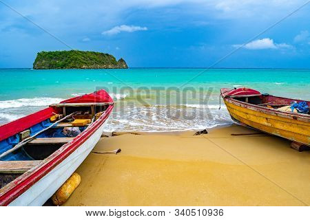 Colorful old wooden fishing boats docked by water on a beautiful beach coast. White sand sea shore landscape on tropical Caribbean island. Holiday weekend/ summer vacation setting at Pagee / Pajee Beach in Port Maria, Jamaica. Cabarita island in the dista