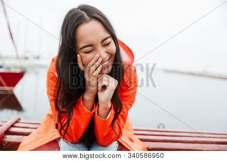 Smiling attractive young asian woman wearing raincoat spending time outdoors walking at the coastland