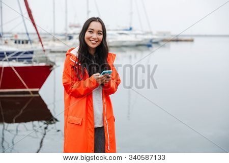 Smiling attractive young asian woman wearing raincoat spending time outdoors walking at the coastland, using mobile phone